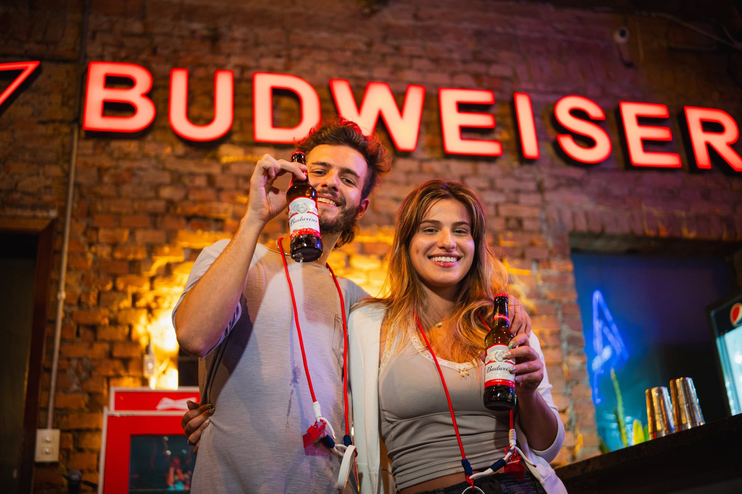 Casal brindando com garrafas de Budweiser diante do letreiro em neon