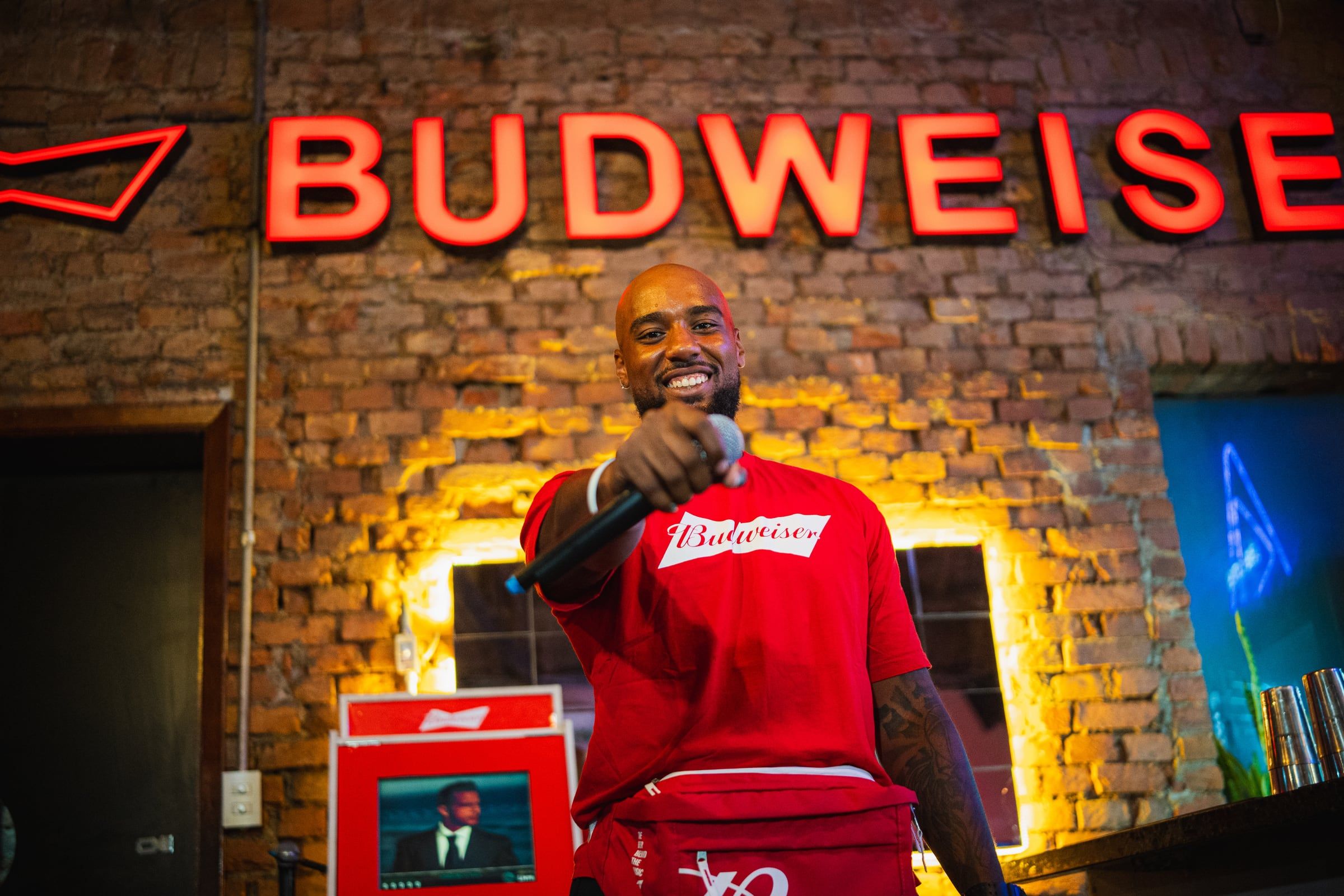 Homem de camiseta da Budweiser apontando para a câmera no palco do bar