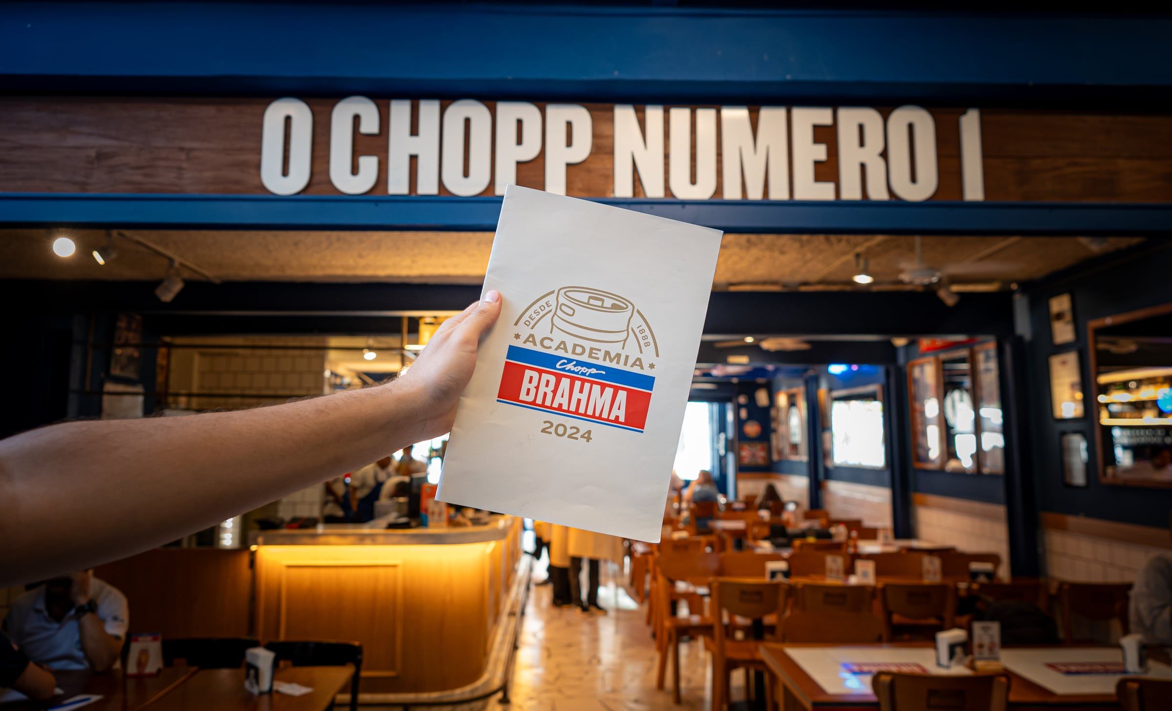 Mão erguendo um impresso do Chopp Brahma no salão do bar, sob a placa O chopp número 1