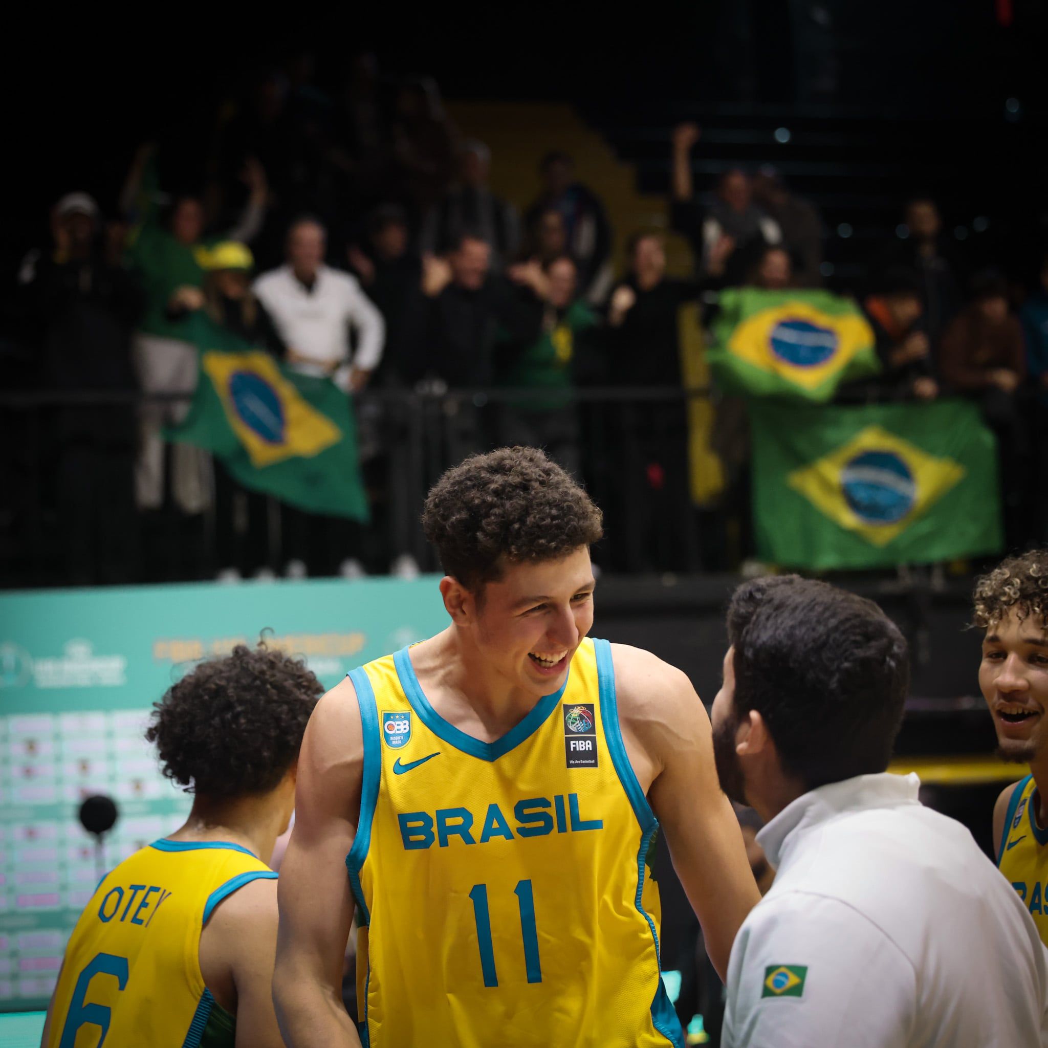 Jogador sorrindo depois do jogo, com torcedores e bandeiras do Brasil ao fundo